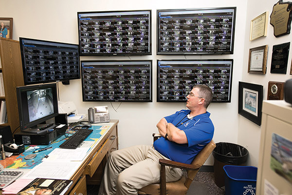 a man sits in front of multiple video monitors displaying surveillance images
