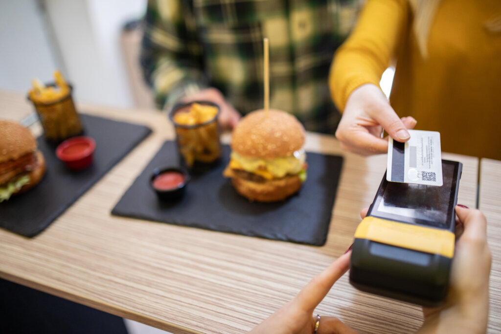 Young woman making contactless payment with credit card for burgers in fast food restaurant, boyfriend standing beside her