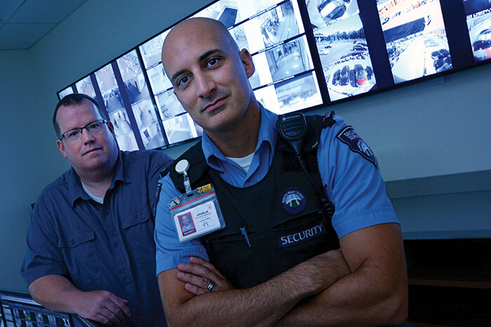 Two security officers stand in the video surveillance monitoring center for Hamilton Health Sciences