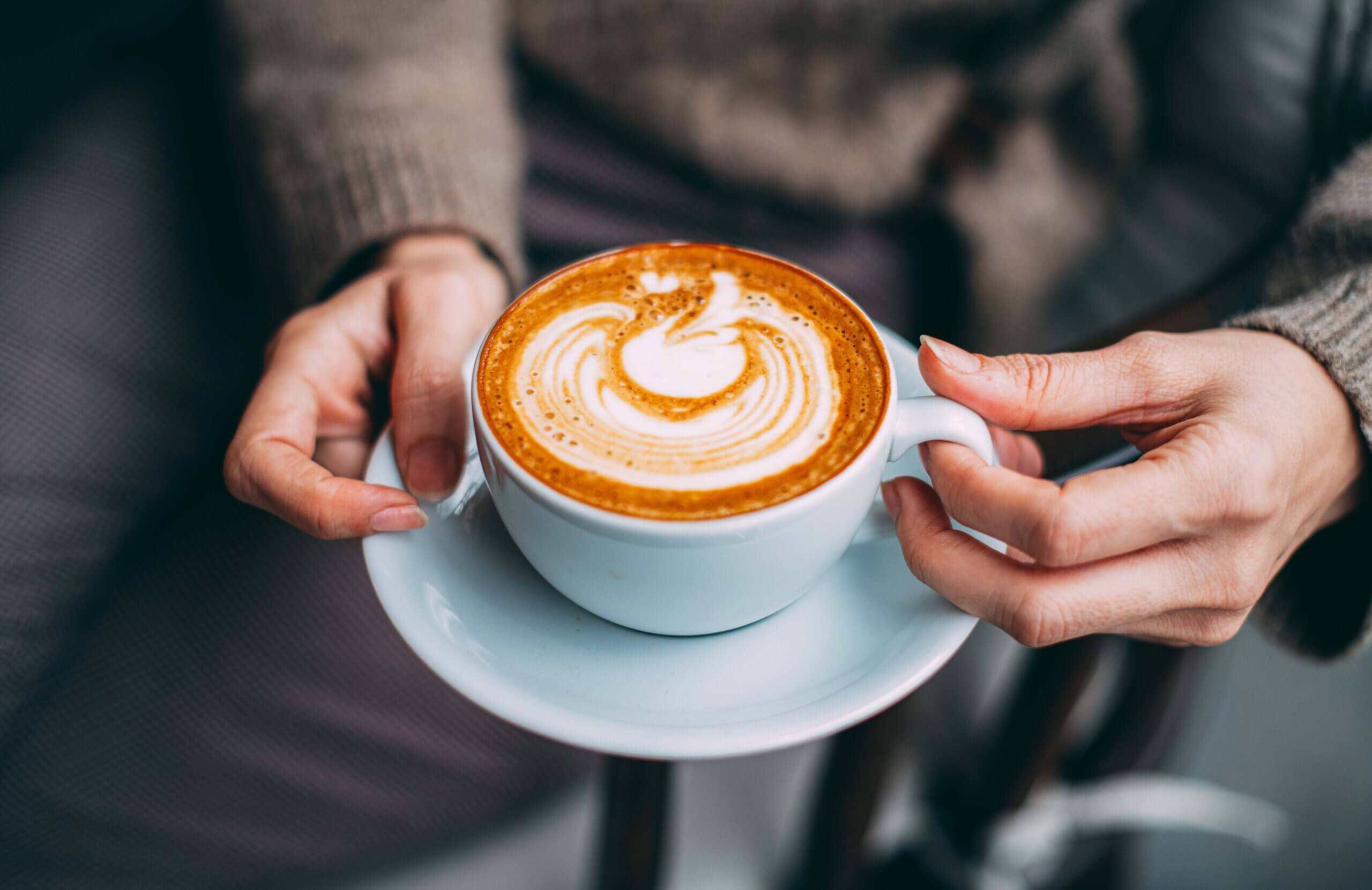 Woman's hands holding frothy latte in white cup and saucer.