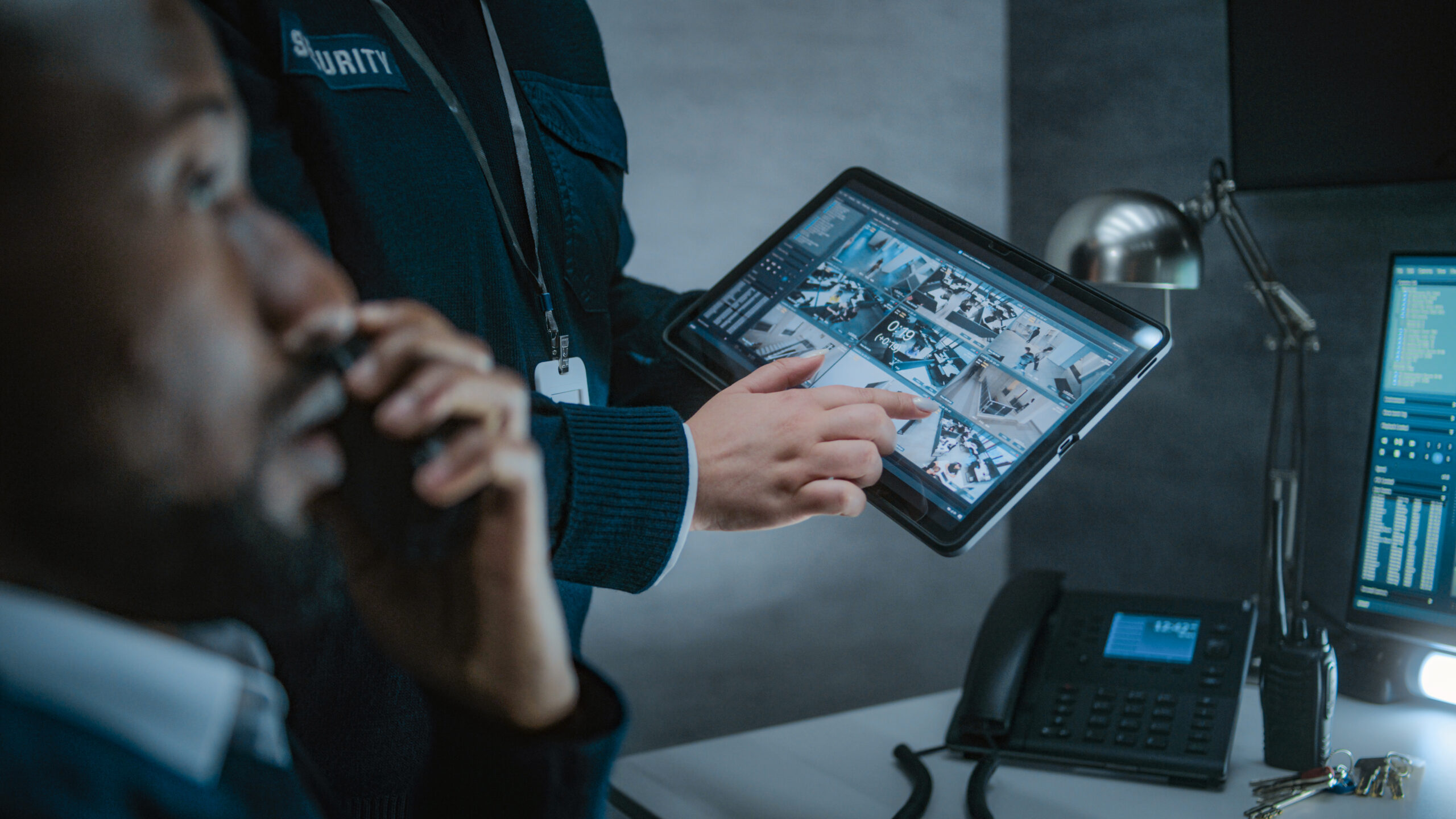 Female employee shows CCTV footage to African American male security officer using tablet computer. Diverse security operators control surveillance cameras on computer monitors in monitoring center.