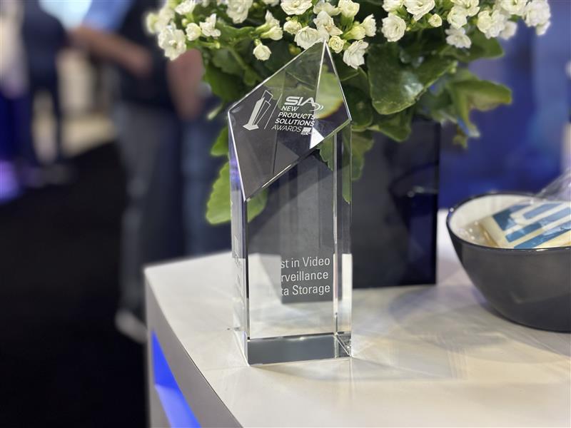 Close-up of a clear glass SIA New Product Solutions Award trophy won by March Networks, displayed on a white tabletop, with small white flowers and a softly blurred trade show background.