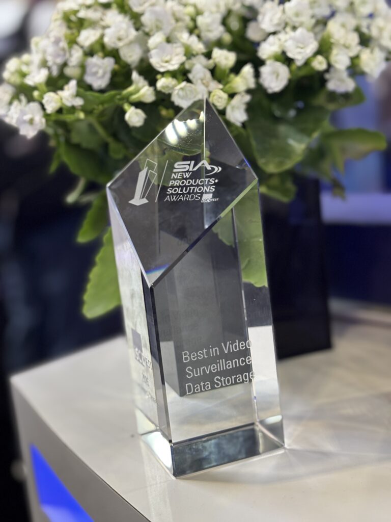 Close-up of a clear glass SIA New Product Solutions Award trophy won by March Networks, displayed on a white tabletop, with small white flowers and a softly blurred trade show background.