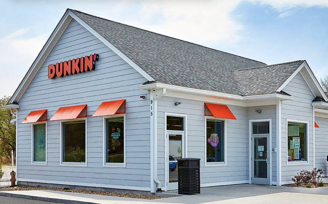 Extérieur d'un restaurant Dunkin' gris, avec des auvents orange aux fenêtres, une entrée principale et un ciel bleu limpide.