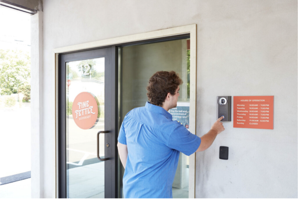 Un homme vêtu d'une chemise bleue utilise un interphone à écran tactile situé à côté d'une porte vitrée portant l'inscription « Entrée du Fine Fettle Dispensary ».